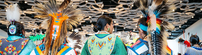 the backs of five indigenous hoop dancers in ceremonial costumes with beads and feathers